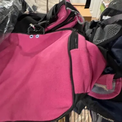 Bright pink canvas duffel bag with black trim and zippers, shown piled with other luggage on a wire shelf.