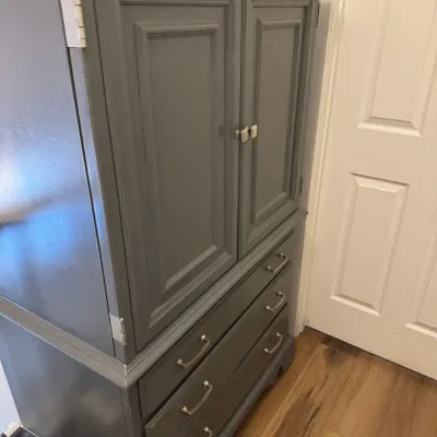 Sale Photo Thumbnail #178: Traditional wooden armoire painted in a modern slate gray, featuring a double-door upper cabinet and three lower drawers with silver-tone hardware.