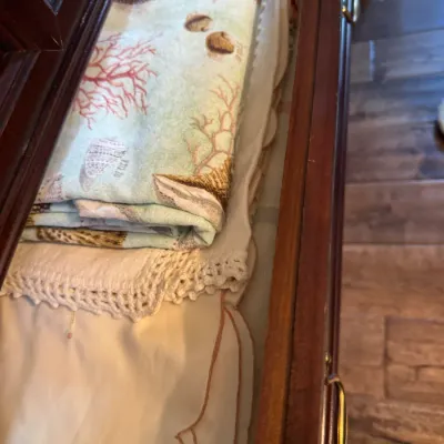 Assorted vintage linens, including a seashell-patterned cloth and a white textile with crocheted lace trim, stored inside a wooden drawer.