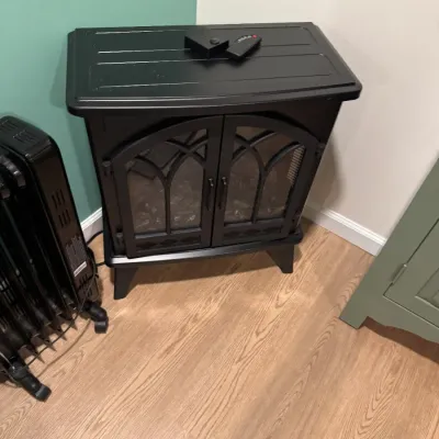 Black electric fireplace stove heater with arched glass doors and a remote control, shown alongside a black oil-filled radiator heater.