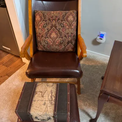 Sale Photo Thumbnail #260: Vintage-style high-back armchair with brown leather upholstery, nailhead trim, and a matching ottoman, paired with a decorative paisley throw pillow and a small map-print trunk.