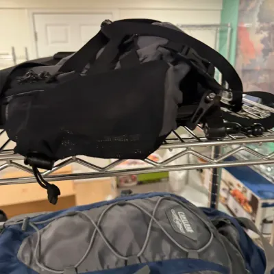 CamelBak hydration pack in grey and blue, shown alongside another black and grey outdoor backpack on a wire shelf.