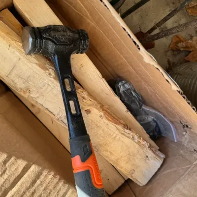 Sale Photo Thumbnail #204: Roughneck ball-peen hammer with a black and orange rubberized grip, resting on wooden blocks inside a cardboard box.
