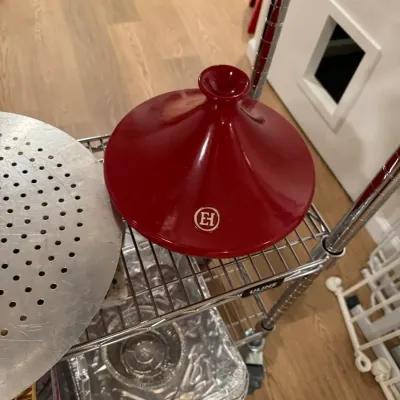 Emile Henry red ceramic tagine lid, showing the brand's signature logo on the side, displayed on a wire shelving unit.