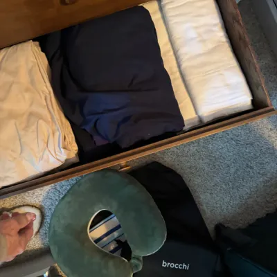 Sale Photo Thumbnail #148: Assorted bed linens folded inside a wooden dresser drawer, shown alongside a green travel neck pillow and a black Brocchi bag on the floor.