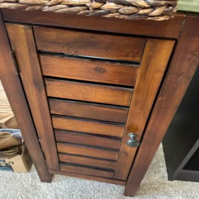 Sale Photo Thumbnail #267: Small wooden accent cabinet with a slatted shutter-style door and a dark metal knob, topped with a woven basket.