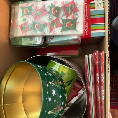 Sale Photo Thumbnail #230: Cardboard box filled with assorted Christmas-themed items, including decorative tins, gift boxes, and holiday napkins.
