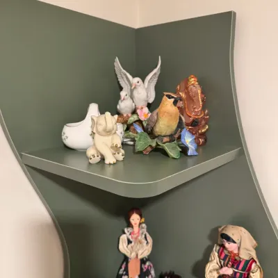 Sale Photo Thumbnail #132: Collection of decorative figurines, including a small elephant, birds, a Ganesha statue, and traditional dolls, displayed on a sage green corner shelf.
