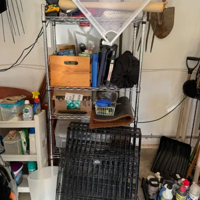 Sale Photo Thumbnail #211: Four-tier chrome wire shelving unit holding various garage items, including a wooden crate, cleaning supplies, and a folded black wire pet crate leaning against the front.