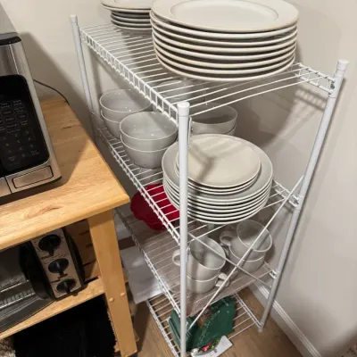Sale Photo Thumbnail #109: White wire four-tier shelving unit holding a collection of neutral-toned ceramic dinnerware, including plates, bowls, and mugs.