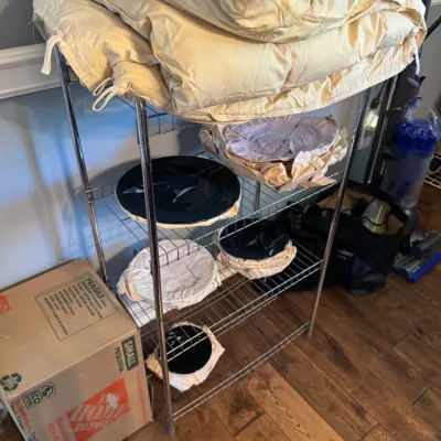 Sale Photo Thumbnail #226: Four-tier chrome wire shelving unit holding a folded comforter and wrapped ceramic plates.