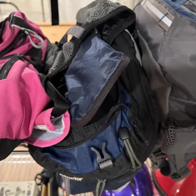 Assorted outdoor backpacks and gear, including a blue and black hydration pack, a pink bag, and a grey backpack, displayed on a wire shelf.