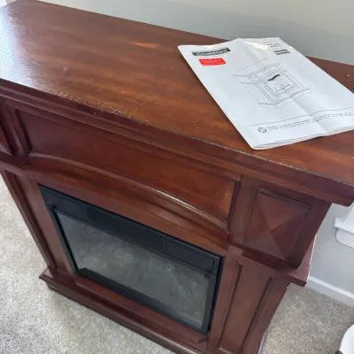 Style Selections electric fireplace with a cherry-finished wooden mantel, showing some light wear on the top edge, complete with the...