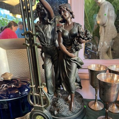 Vintage dark bronze-finished figural clock statue featuring a romantic couple, shown alongside silver-plated goblets and a cobalt blue...