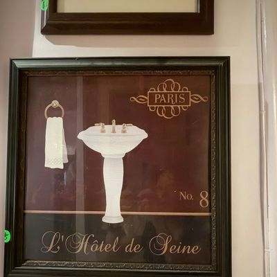 Framed Paris-themed bathroom wall art featuring a pedestal sink, towel illustration, and French typography in a dark moulded frame.