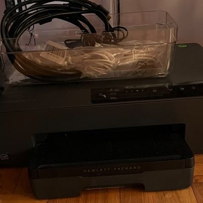 Black Hewlett-Packard all-in-one printer with an attached output tray and a clear plastic bin containing power and connection cables...