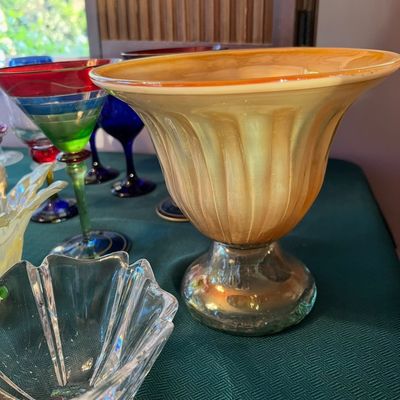 Large iridescent amber-toned art glass pedestal vase featuring ribbed detailing, surrounded by colorful glassware.