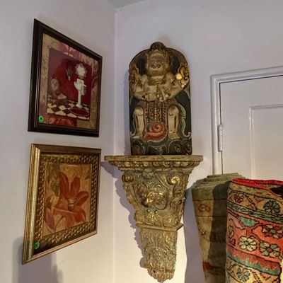 Hand-carved Balinese deity statue displayed atop a gilded ornate wall sconce shelf, flanked by framed artwork and rolled area rugs.