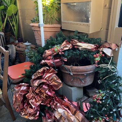 Large artificial holiday wreath decorated with metallic bronze bows, string lights, and berries, positioned outdoors near terracotta...