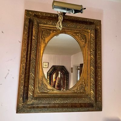 Ornate antique giltwood framed wall mirror with an oval center pane, fitted with a brass picture light overhead.