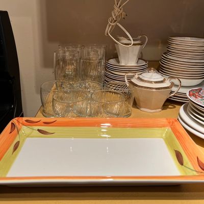 Hand-painted rectangular ceramic serving platter with an orange and green leaf border, alongside stacks of porcelain dinnerware and clear...