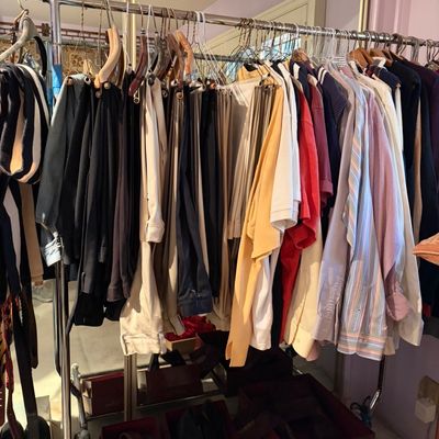 A metal rolling garment rack filled with a variety of men's and women's clothing, including trousers, button-down shirts,...