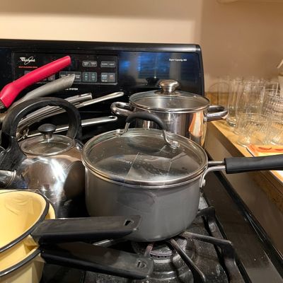 Collection of assorted kitchen cookware on a stovetop, featuring a grey Cuisinart saucepan with a glass lid, stainless steel stockpot,...