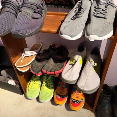 A collection of modern footwear arranged on a wooden shoe rack, including athletic sneakers, toe shoes, flip-flops, and slip-on casual...