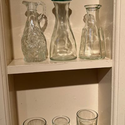 Collection of vintage clear glassware displayed on shelves, including press glass carafes, a decanter, and drinking glasses.
