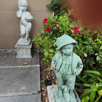 Two vintage concrete garden statues depicting young boys, including one on a square pedestal holding a basket and one in alpine-style...