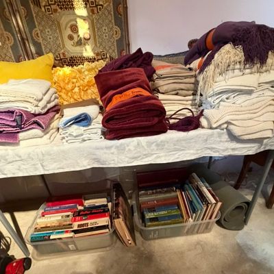 Folded household linens, towels, and throw pillows arranged on a table, with plastic bins of vintage books stored underneath.