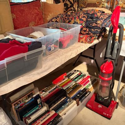 Red Dirt Devil upright vacuum cleaner alongside clear storage tubs filled with linens, clothing, and hardcover books.