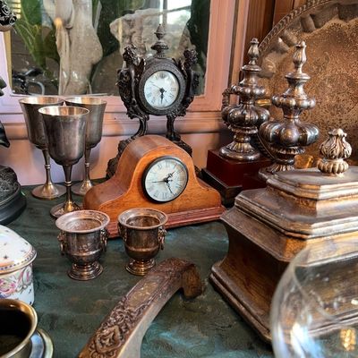 Collection of vintage decor items including an ornate metal desk clock, a wooden mantle clock, tarnished silver goblets, and decorative...
