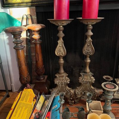 Collection of tall candlesticks including carved wooden floor stands and ornate brass pedestal holders topped with red pillar candles,...