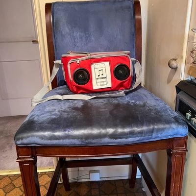 Red soft-sided cooler bag with built-in portable speakers and shoulder strap, resting on a blue upholstered wooden dining chair.