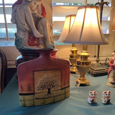 Flat ceramic vase featuring a textured landscape and tree design, displayed alongside brass table lamps and Asian porcelain figurines.