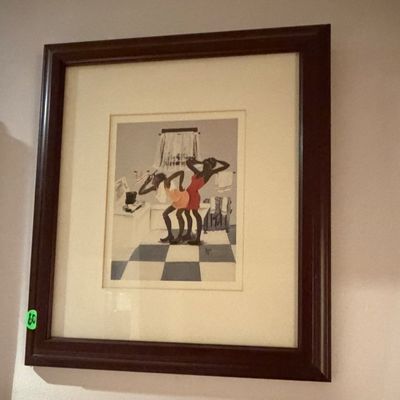 Framed artwork depicting figures in a bathroom setting, presented with wide matting in a dark wood frame.