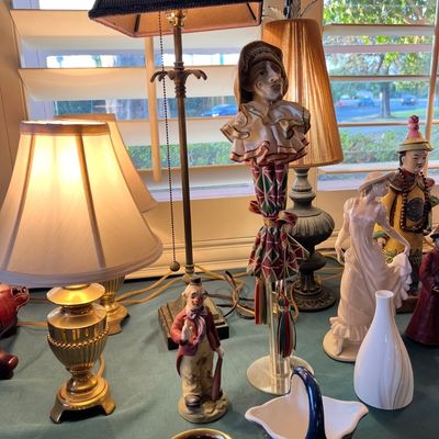 Collection of table lamps, porcelain figurines including clowns and a lady in a dress, and decorative ceramic items displayed on a tabletop.
