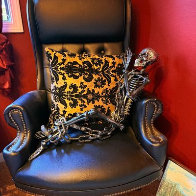 Sale Photo Thumbnail #3: High-back black leather executive office chair with brass nailhead trim, paired with a gold and black damask accent pillow and a decorative Halloween skeleton.