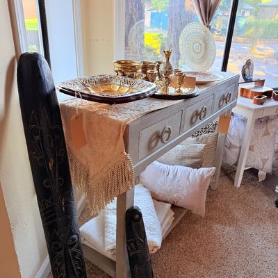 Sale Photo Thumbnail #8: Contemporary gray console table with drawers, styled with brass and silver-toned decorative tableware, flanked by two tall, dark-stained carved wooden African-style masks.