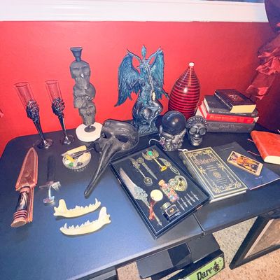 Sale Photo Thumbnail #4: Assortment of gothic and occult-themed decor, including a Baphomet statue, skull-themed goblets, a plague doctor mask, a quill writing set, and various books on witchcraft.