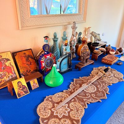 Sale Photo Thumbnail #7: Assorted vintage collectibles displayed on a blue tablecloth, including religious icons, decorative ceramic decanters, a carved elephant figurine, and a ceremonial sword resting on a beaded table runner.