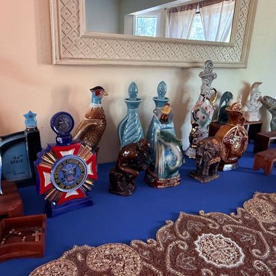 Sale Photo Thumbnail #6: Collection of vintage ceramic decanters, including a Veterans of Foreign Wars commemorative bottle, a pheasant, and various figural designs, displayed alongside a beaded table runner.