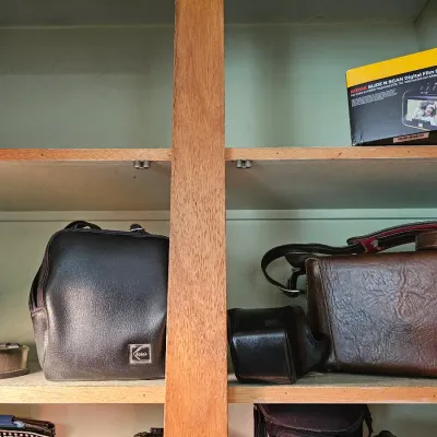Vintage camera bags, including a black Kodak bag and a brown Marsand bag, displayed on wooden shelves alongside a boxed Kodak Slide N...