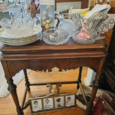 Vintage wooden side table with turned legs and a decorative carved apron, displaying an assortment of cut glass bowls, plates, and a...
