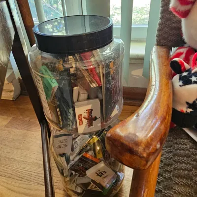 Two large clear plastic jars filled with a collection of vintage matchbooks, displayed on the floor next to a wooden chair.