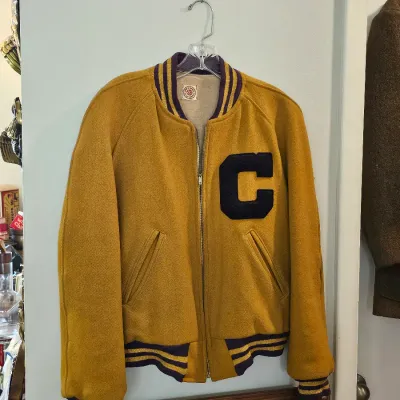 Vintage mustard yellow wool varsity jacket with a purple and yellow striped collar, cuffs, and waistband, featuring a large purple...