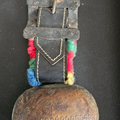 Vintage Swiss-style cowbell featuring a rustic, weathered metal bell attached to a black leather strap with colorful wool trim and a...