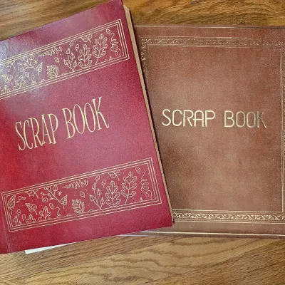 Two vintage scrapbooks with faux leather covers, one in burgundy with a cord tie and gold leaf-patterned borders, and the other in brown...