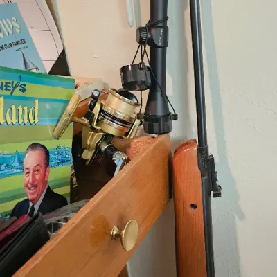 Vintage air rifle with a wooden stock, shown alongside a rifle scope and a gold-colored spinning fishing reel.